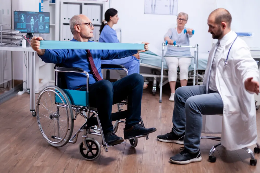 Doctor guiding an older man in a wheelchair through resistance band exercises in a therapy room