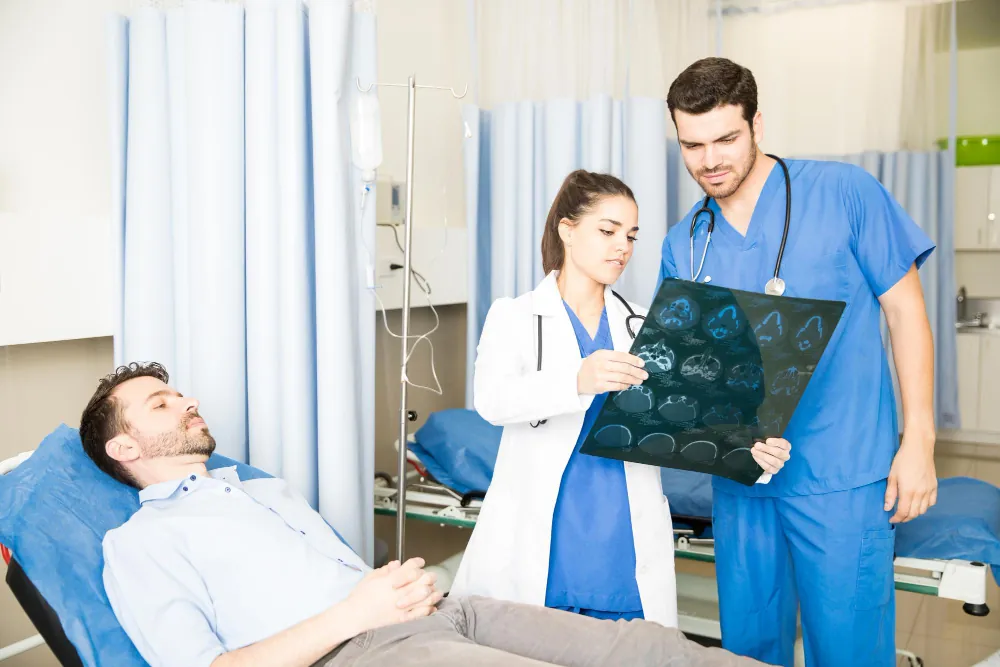 Two doctors reviewing scan films beside a patient resting in a hospital bed